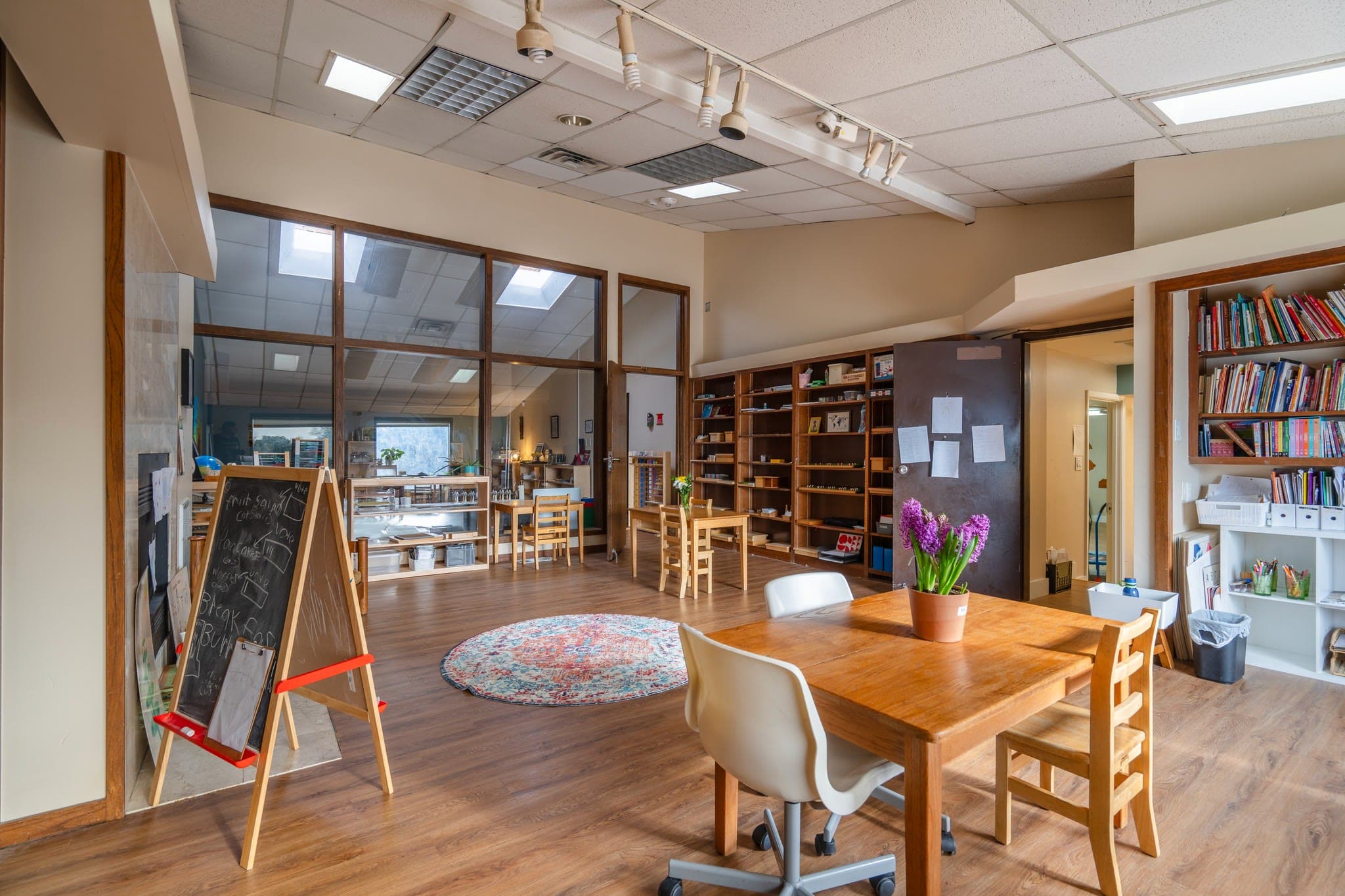 A primary classroom with low shelves of Montessori materials, a chalkboard easel, and a round rug