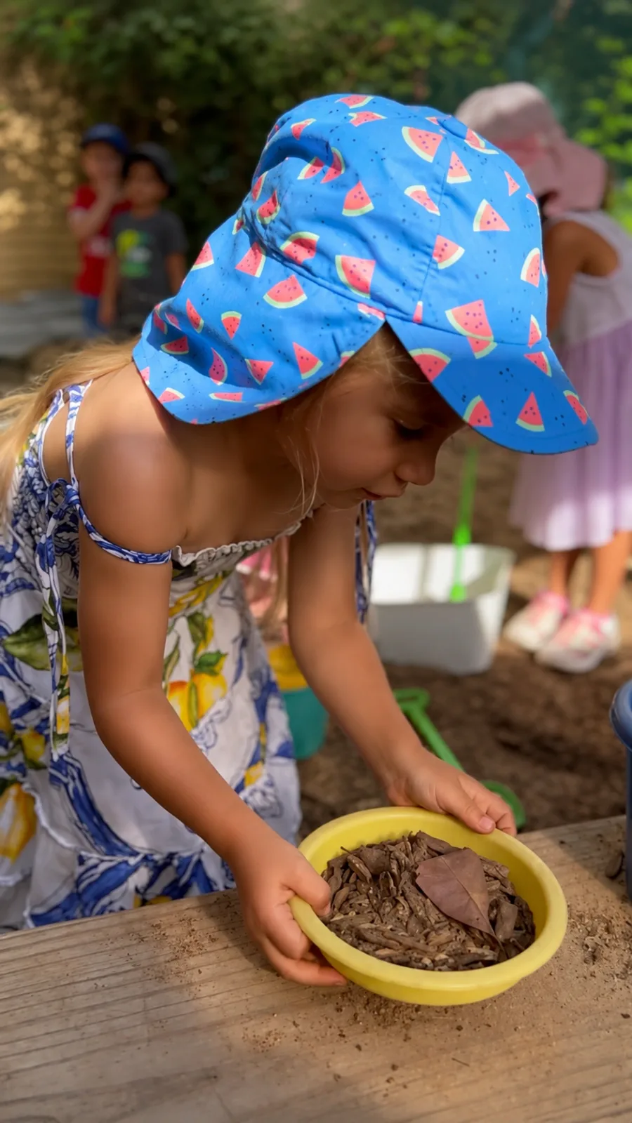 A child in a sun hat exploring a bowl of mulch and leaves at the sand table